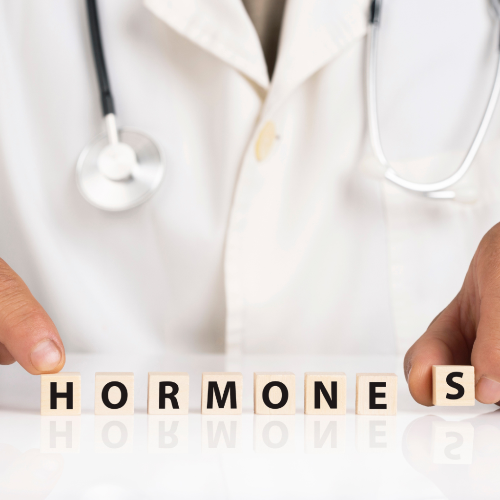 The image shows a person in a white lab coat with a stethoscope around their neck. Wooden blocks at the bottom spell out the word 'HORMONES.'