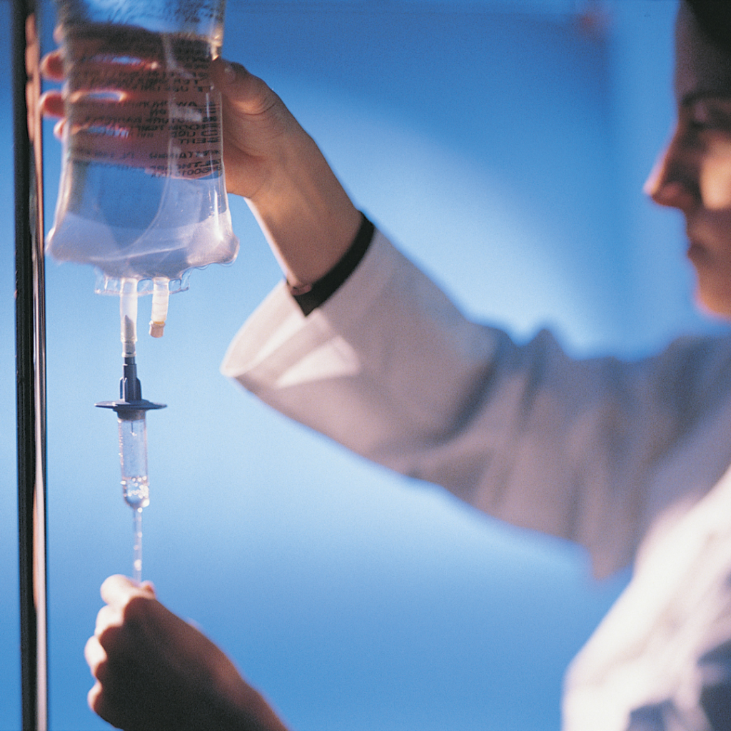 The image shows a close-up of a medical professional adjusting an IV drip. A clear intravenous (IV) fluid bag hangs on a metal stand, with tubing extending downward.