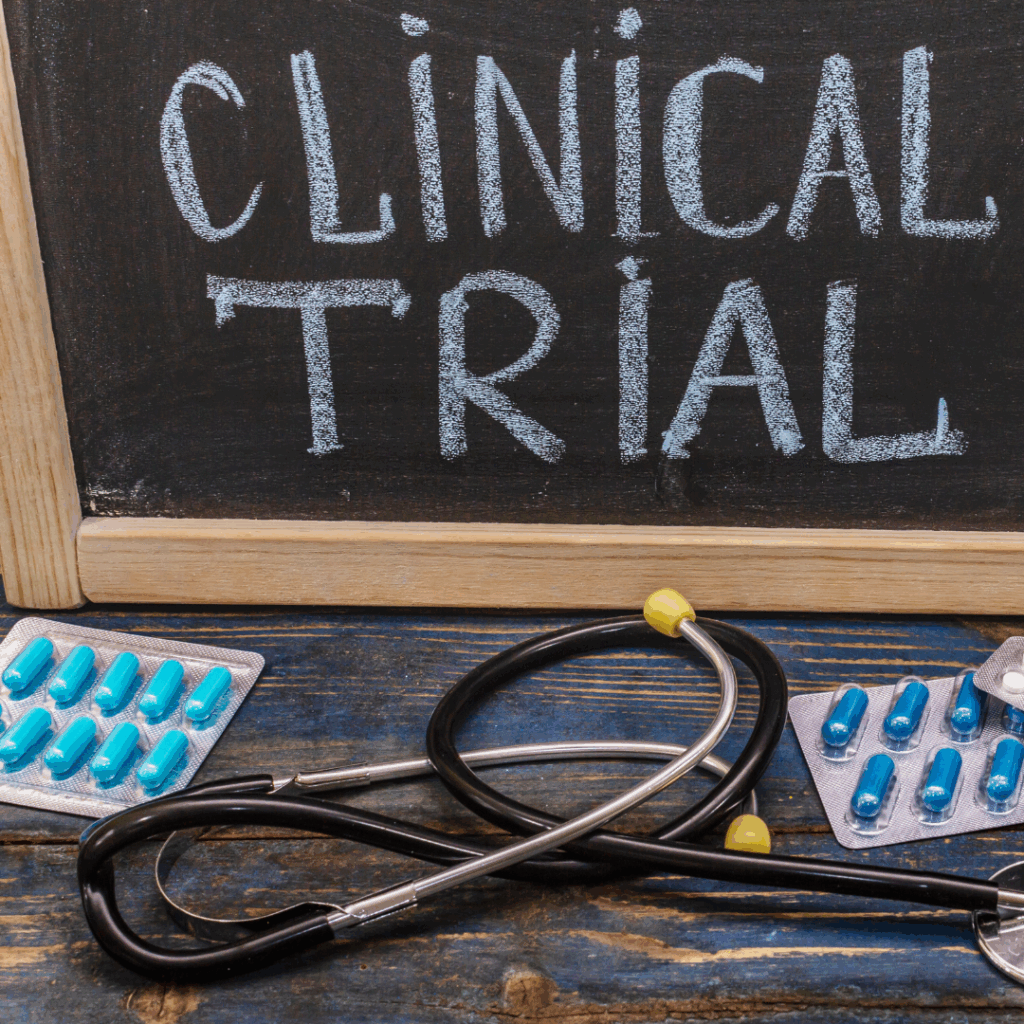 A chalkboard with the words "Clinical Trial" written on it, placed behind a stethoscope and packs of blue capsules on a wooden surface.