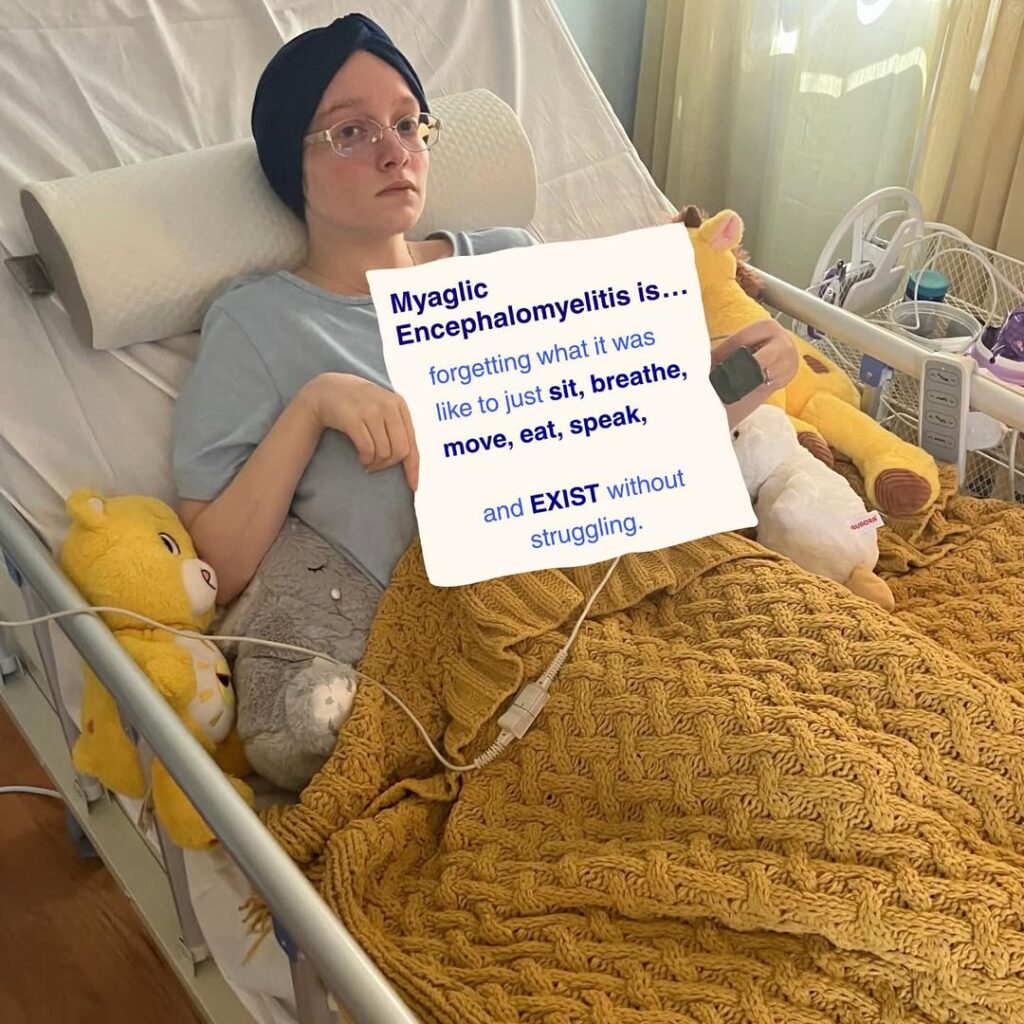 Em is holding a sign that reads: Myalgic Encephalomyelitis is forgetting what it was like to just sit, breathe, move, eat, speak, and EXIST without struggling.