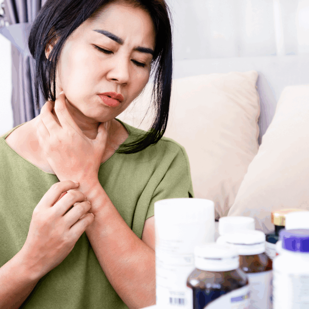 A woman holding her throat and wrist with a pained expression, showing red skin irritation, with various medicine bottles in front of her.