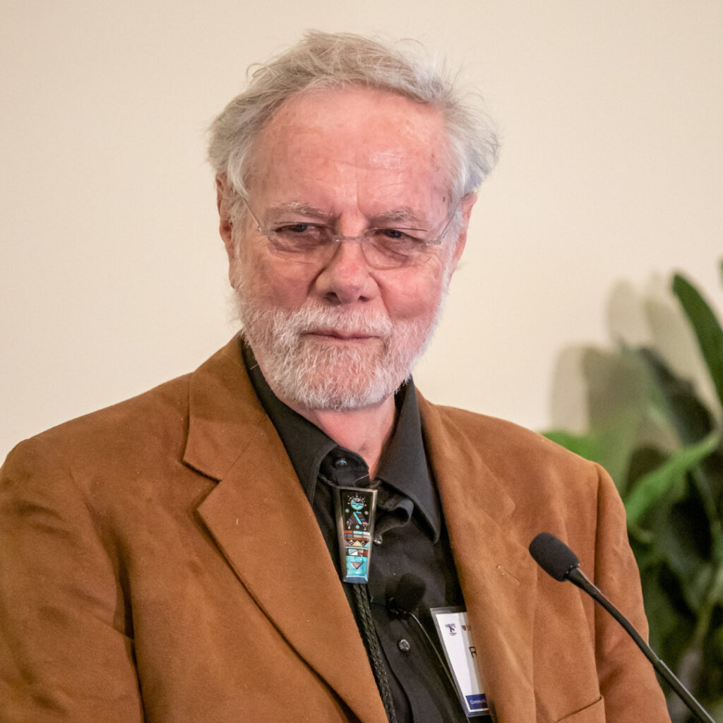 Photo of Ron Davis with gray hair and beard, wearing glasses, a brown blazer, black shirt, and a turquoise bolo tie, speaking at a podium with a lapel microphone.