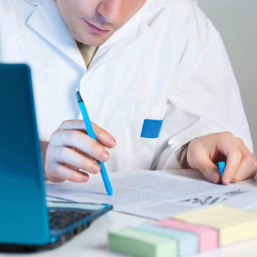 A researcher in a white lab coat reviews a printed scientific paper, highlighting text with a blue pen beside a laptop.